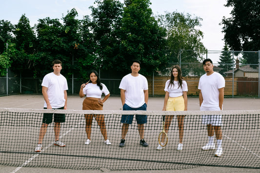 A group of 5 young people on a tennis court, wearing white blank shirts and different colored fleece sweatshorts