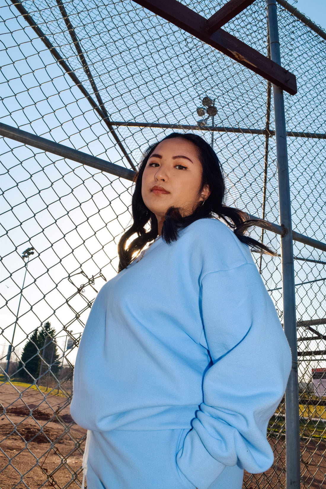 A woman near a baseball field in light blue blank crewneck and sweats